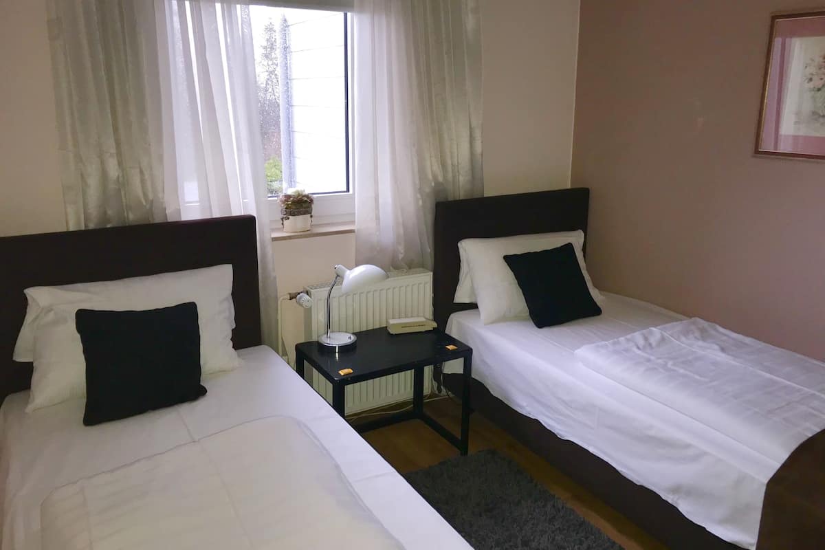 A hotel room with two single beds, a small table with a lamp, a radiator, and a window with curtains.