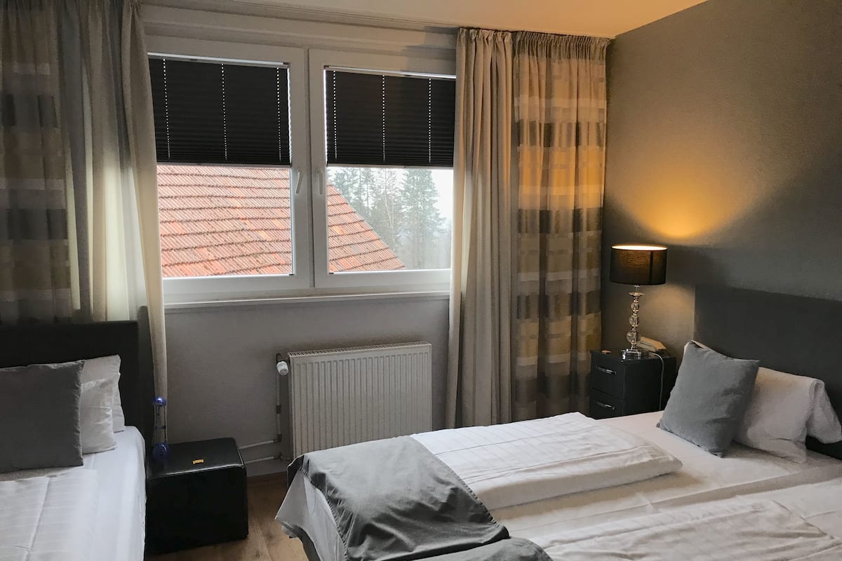 A hotel room with two beds, a radiator, a window with blinds, a nightstand with a lamp, and a view of a red-roofed building outside.