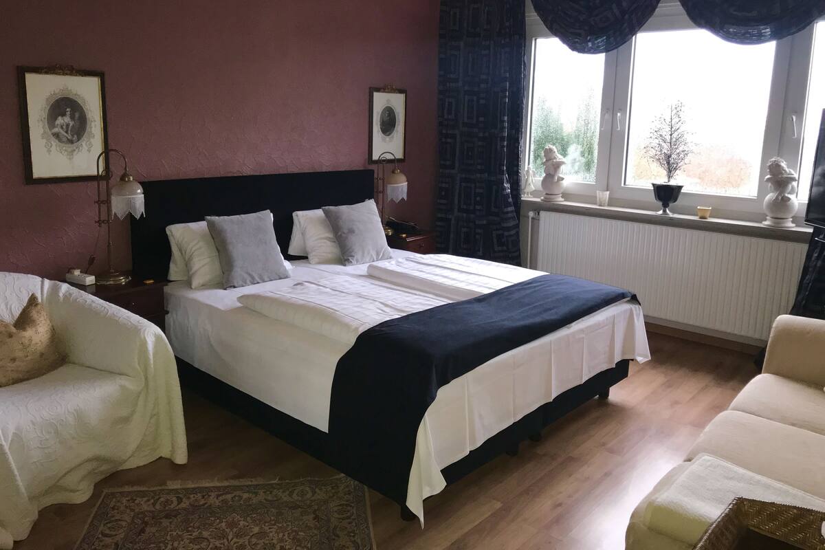 A bedroom with a large bed, two bedside tables, a nightstand, a chair, and a window with curtains.