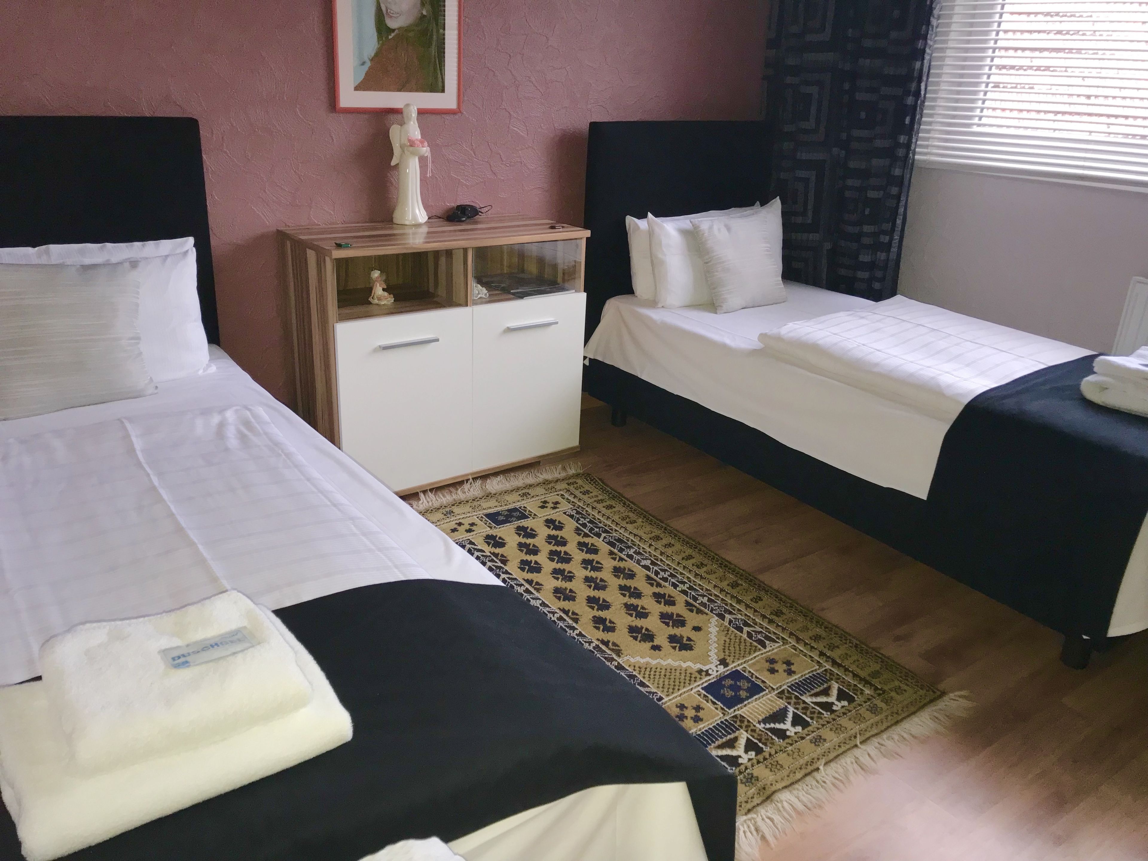Ein Hotelzimmer mit zwei Einzelbetten, einem Holzschrank mit Glastüren, einem gemusterten Teppich und einem Fenster mit Jalousien.