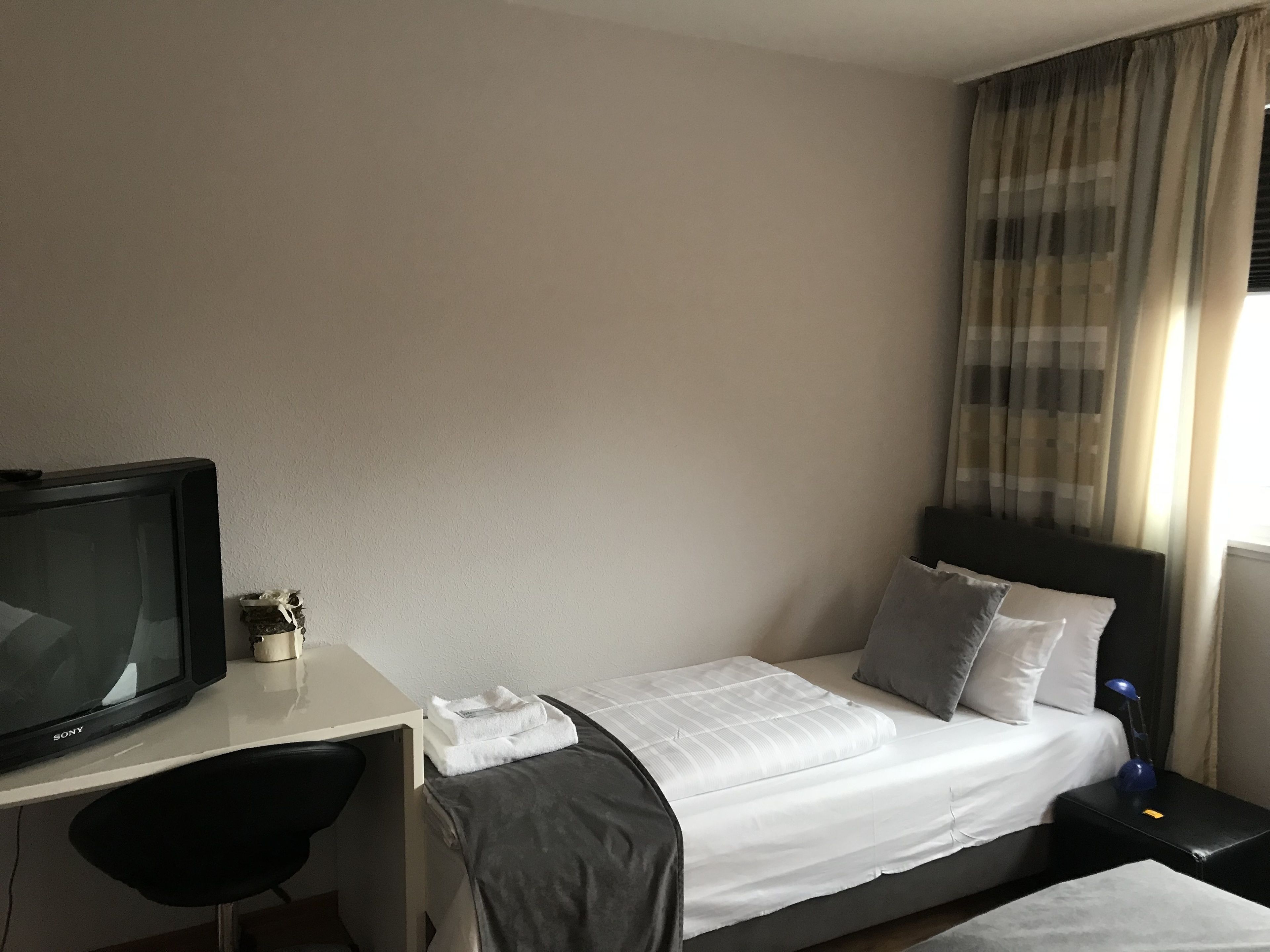 A hotel room with a bed, a desk with a television, a chair, and a nightstand with a lamp.