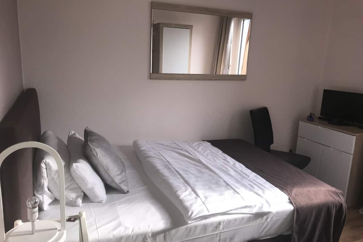 A neatly made bed with white and brown bedding, a bedside table with a lamp, a mirror on the wall, and a TV on a dresser.