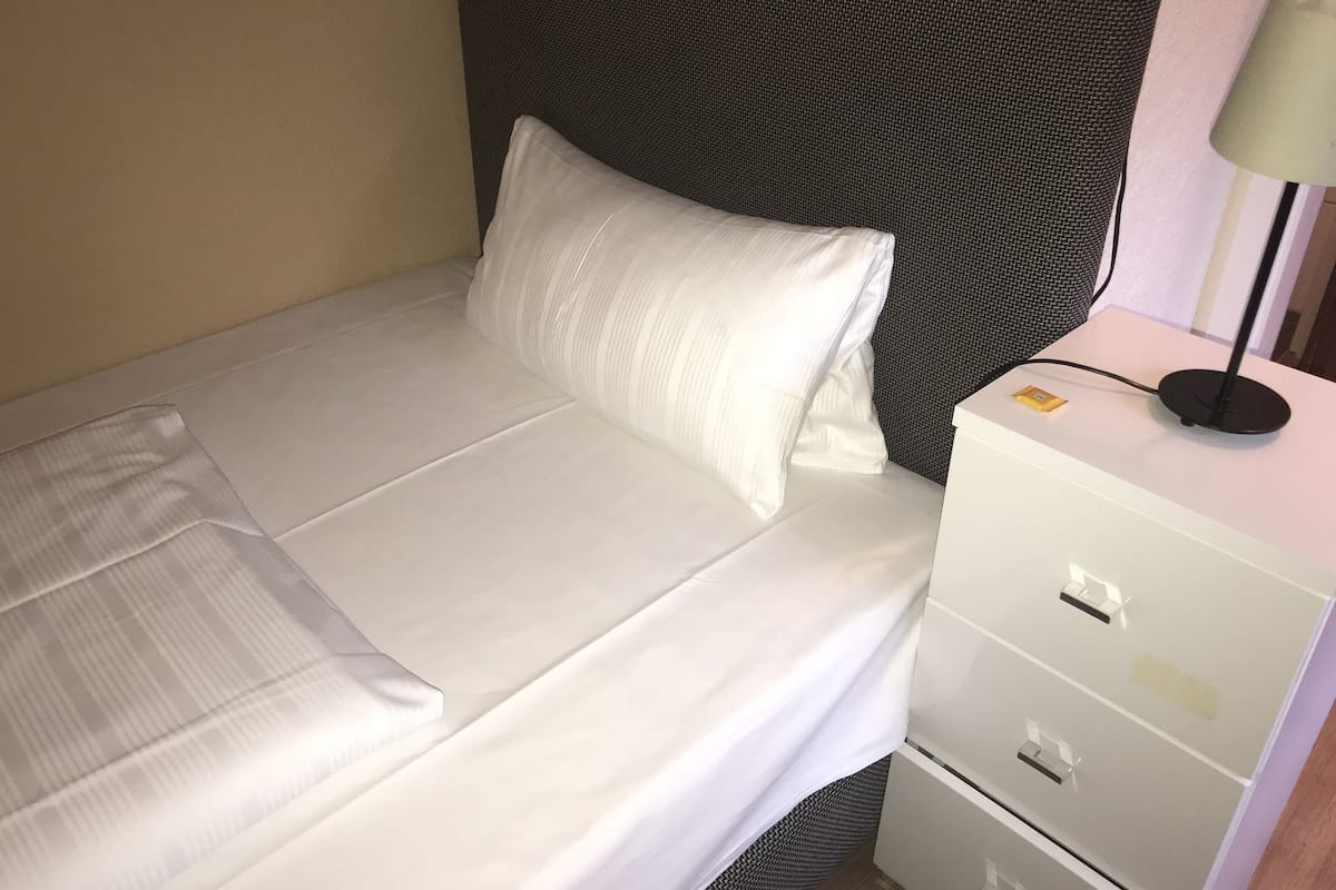 A neatly made bed with white linens, a grey headboard, and a bedside table with drawers.