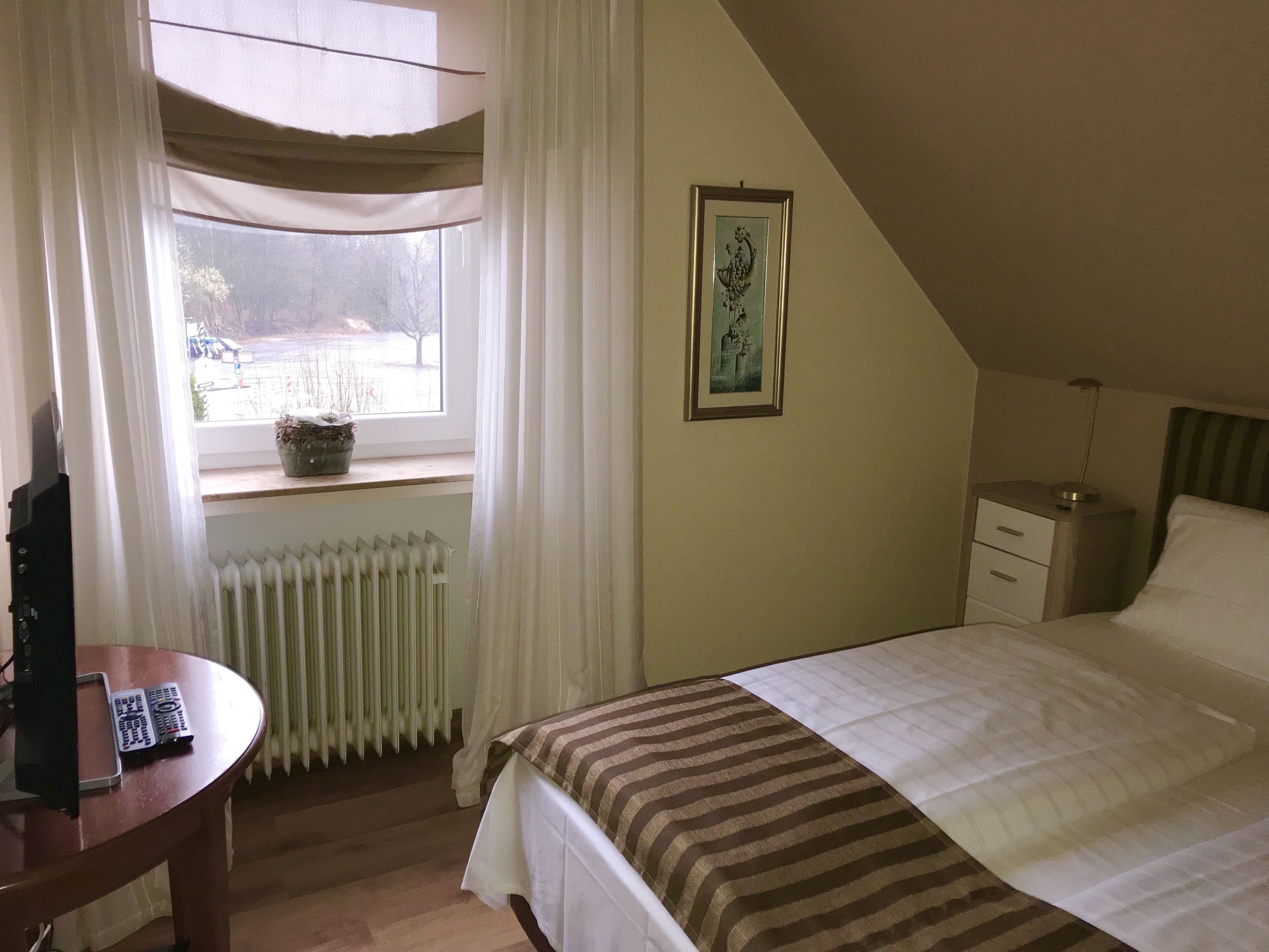 Ein Schlafzimmer mit Bett, Nachttisch, Lampe, einem Fenster mit Vorhängen, einer Heizung und einem gerahmten Bild an der Wand.