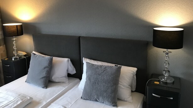 Ein ordentlich bezogenes Bett mit zwei Kissen, ein Nachttisch mit Lampe und eine Wandlampe.