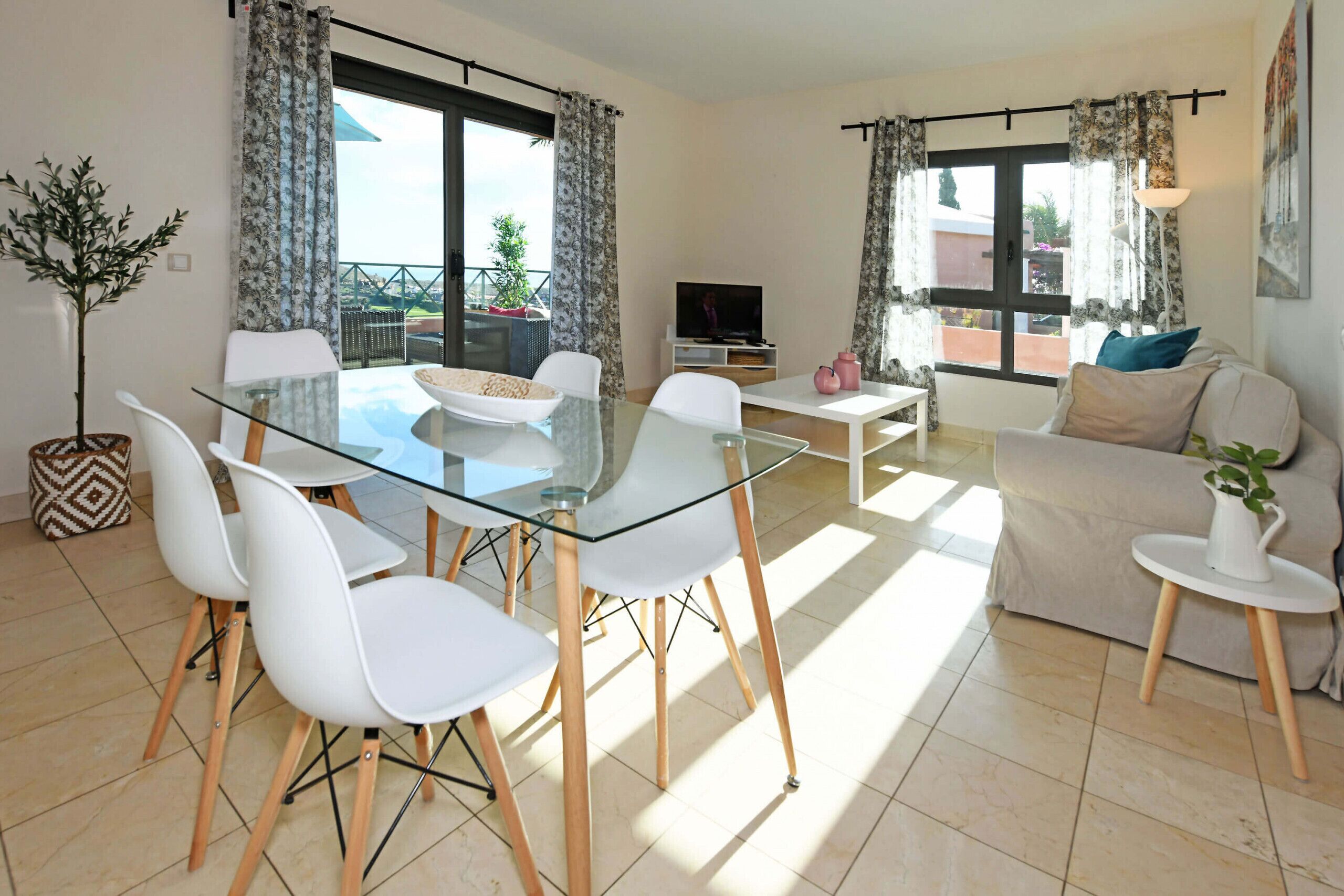 Een moderne woonkamer met een glazen eettafel, witte stoelen, een bank en een tv op een dressoir.