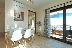 A dining area with a glass table and white chairs, a potted plant, and a view of the sea through a sliding glass door. - Villa Salobre Morro Golf 2 (Maspalomas)