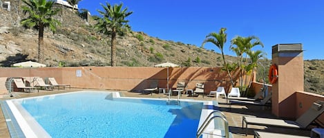 Una piscina con sillas de descanso y palmeras, rodeada por una ladera.
