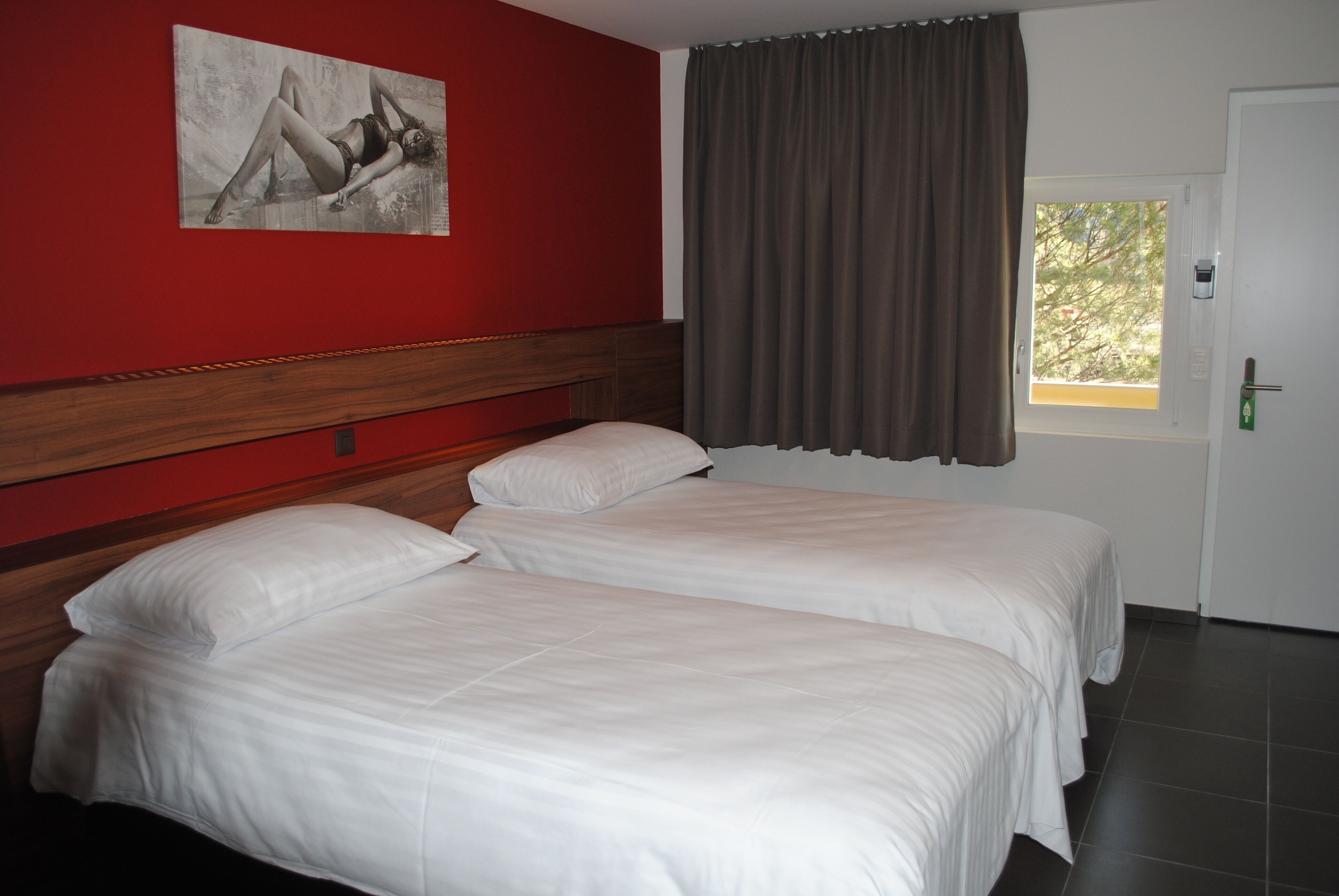 Habitación de hotel con dos camas individuales, un cabecero de madera, una pared de acento roja, una ventana con cortinas y una puerta con cerradura.