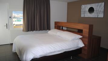 Cama bem arrumada com lençóis brancos em quarto com cabeceira marrom, janela com cortinas e uma obra de arte abstrata na parede.