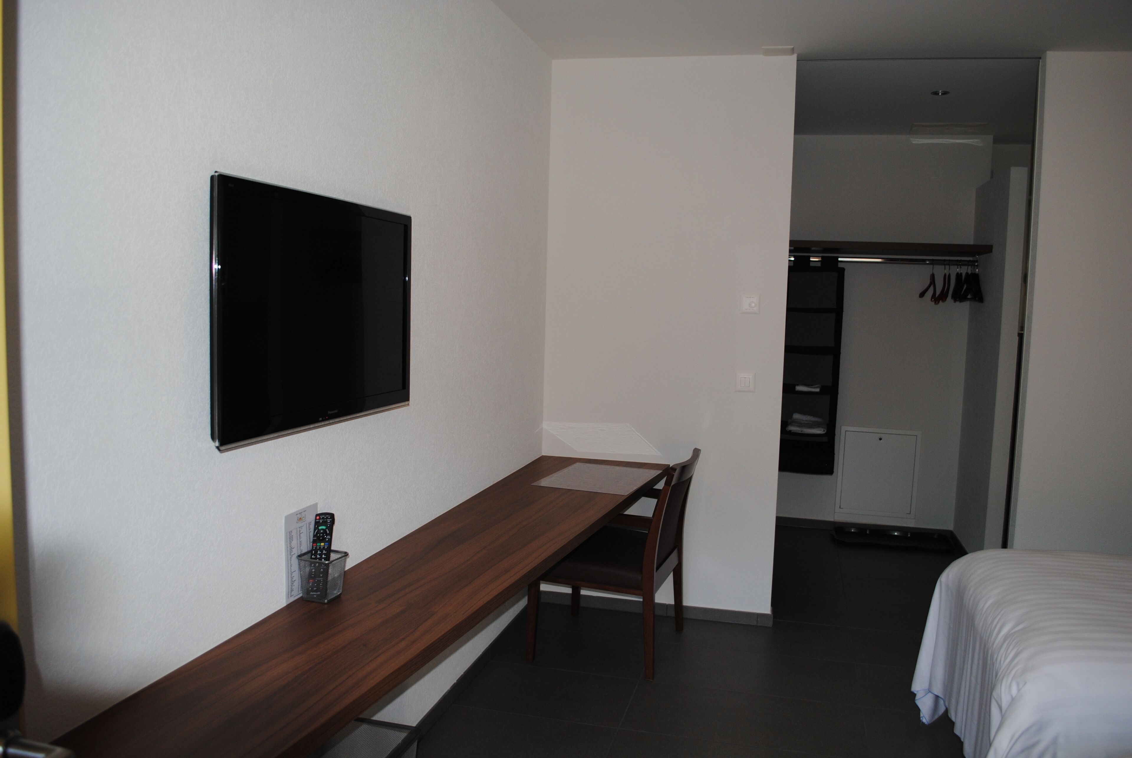 Habitación de hotel con escritorio de madera, televisor montado en la pared, cama y vestidor con ropa colgada.