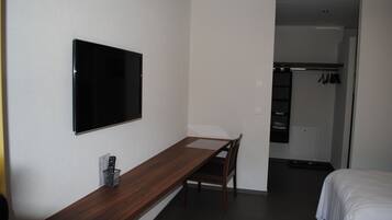 Habitación de hotel con escritorio de madera, televisor montado en la pared, cama y vestidor con ropa colgada.