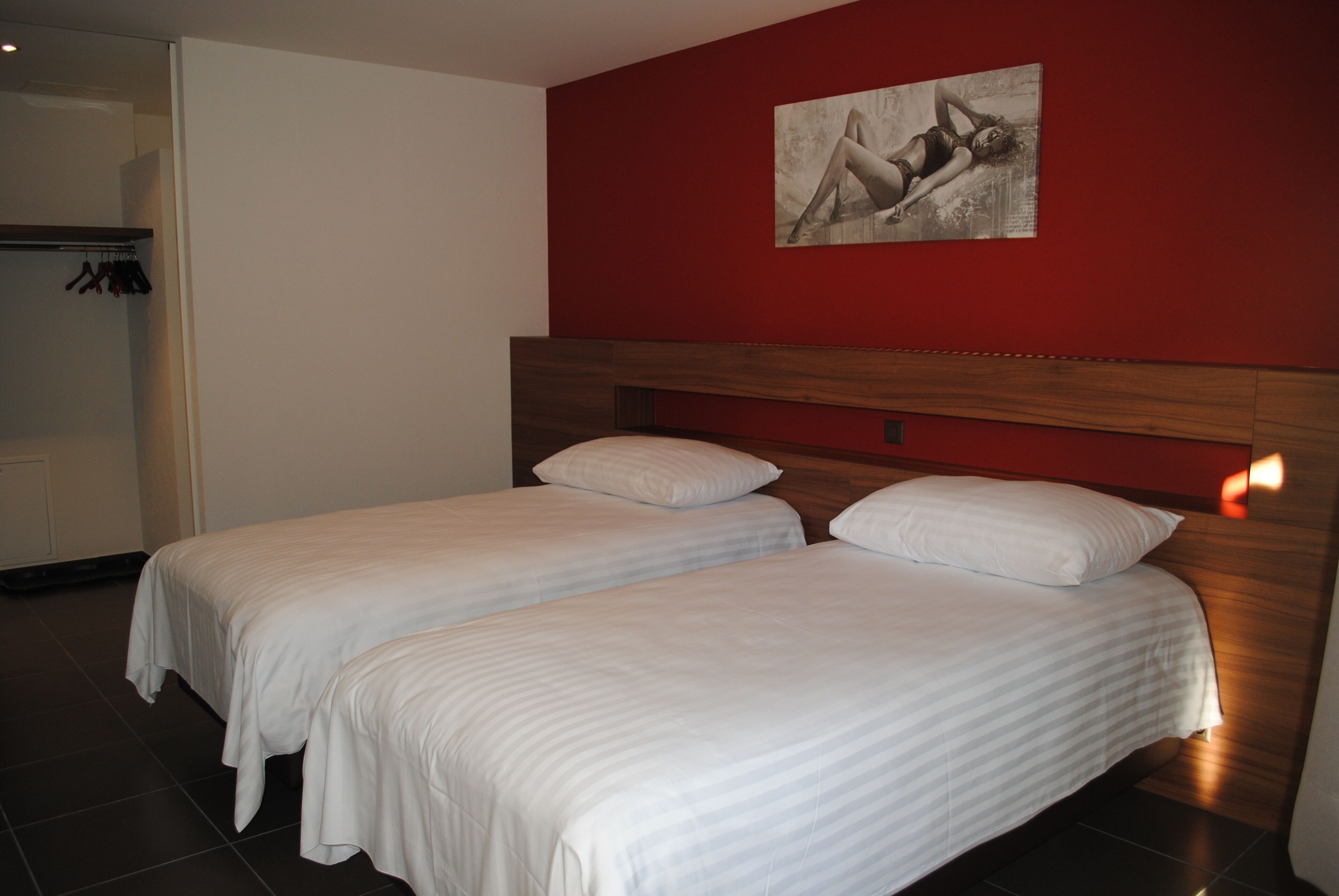 Une chambre d’hôtel avec deux lits simples, une tête de lit en bois, un mur d’accent rouge et un tableau.