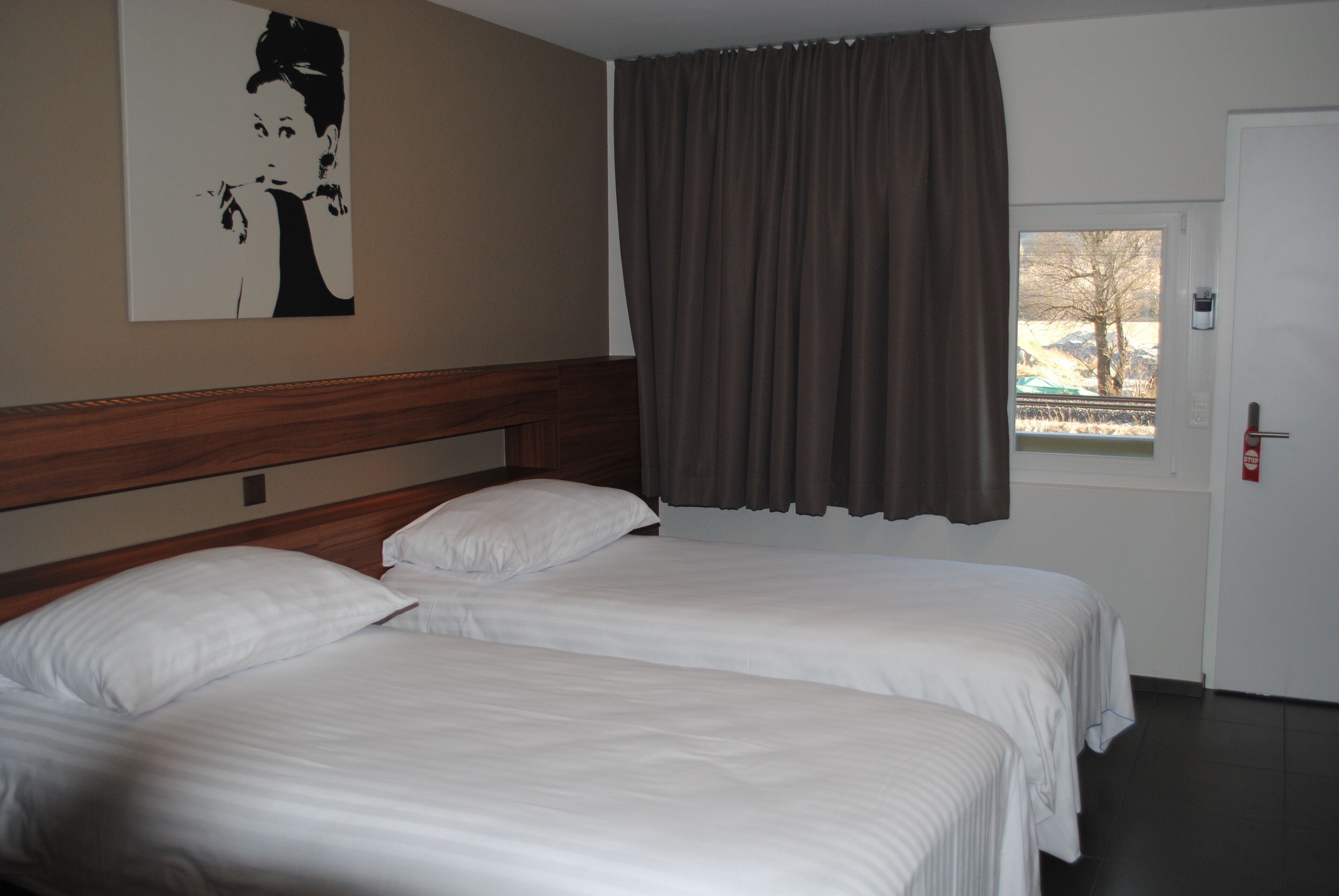 A hotel room with two beds, a wooden headboard, a window with curtains, and a door.