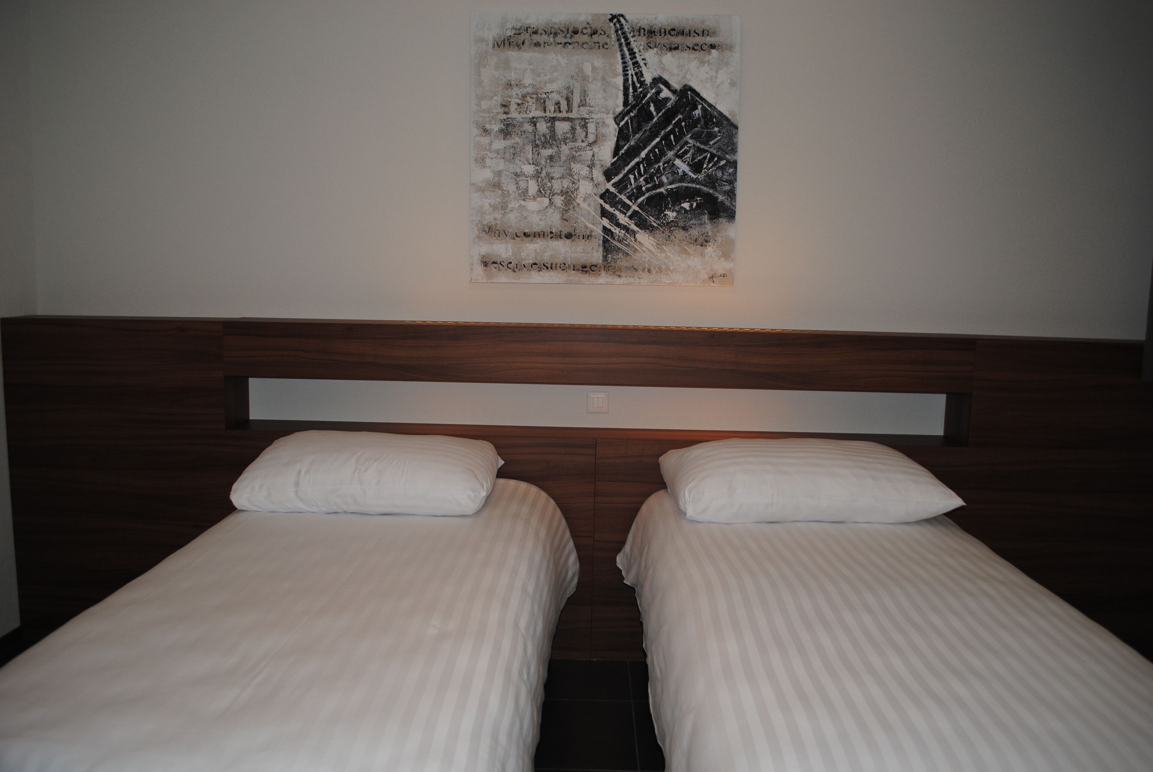 A hotel room with two single beds, a wooden headboard, and a wall-mounted artwork depicting a building.