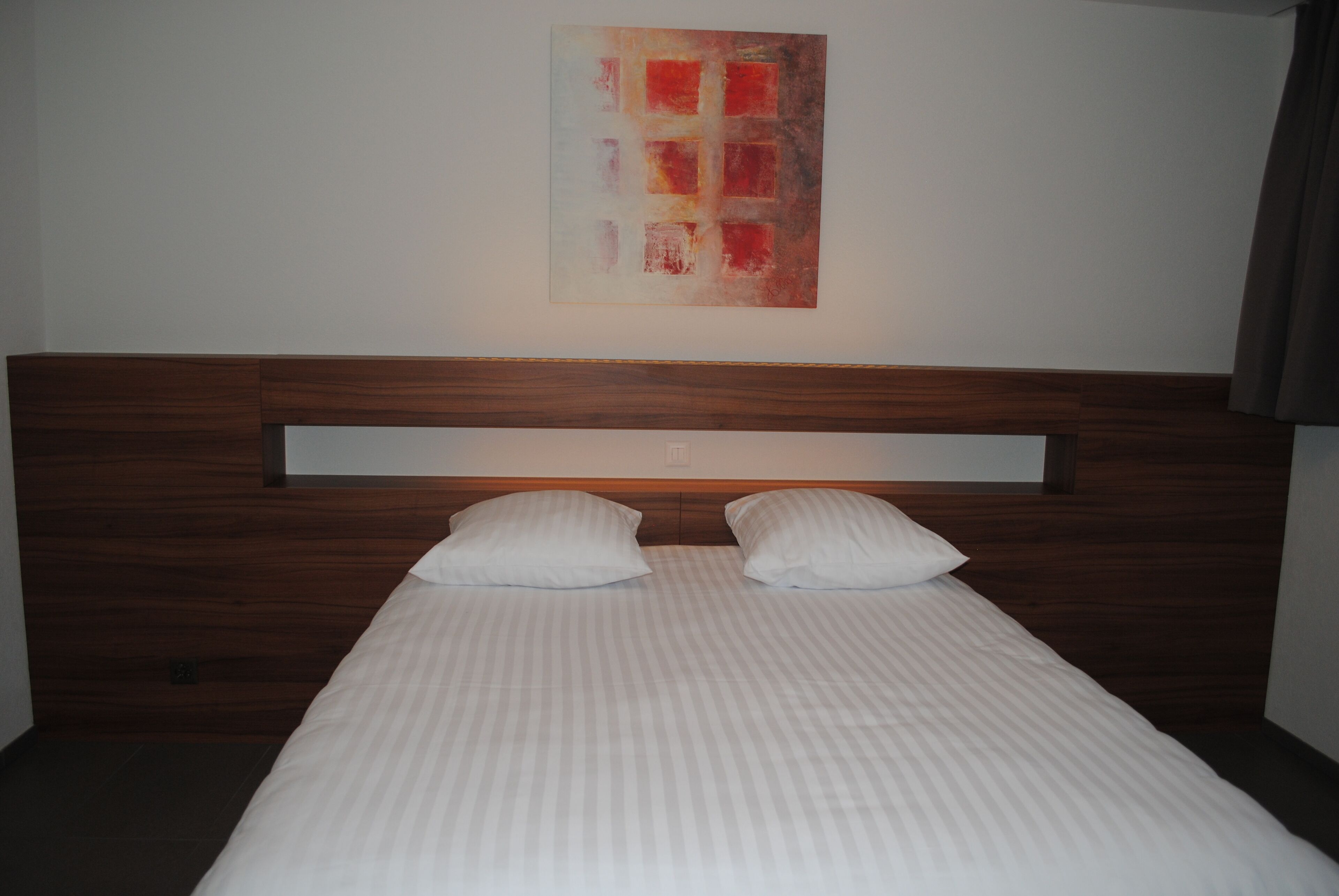Un lit bien fait, avec une tête de lit en bois et deux oreillers, un mur sobre orné d’une seule œuvre d’art abstraite, et une pièce au décor simple et épuré.