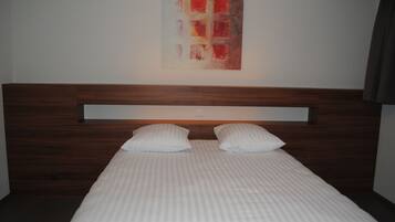Un lit bien fait, avec une tête de lit en bois et deux oreillers, un mur sobre orné d’une seule œuvre d’art abstraite, et une pièce au décor simple et épuré.
