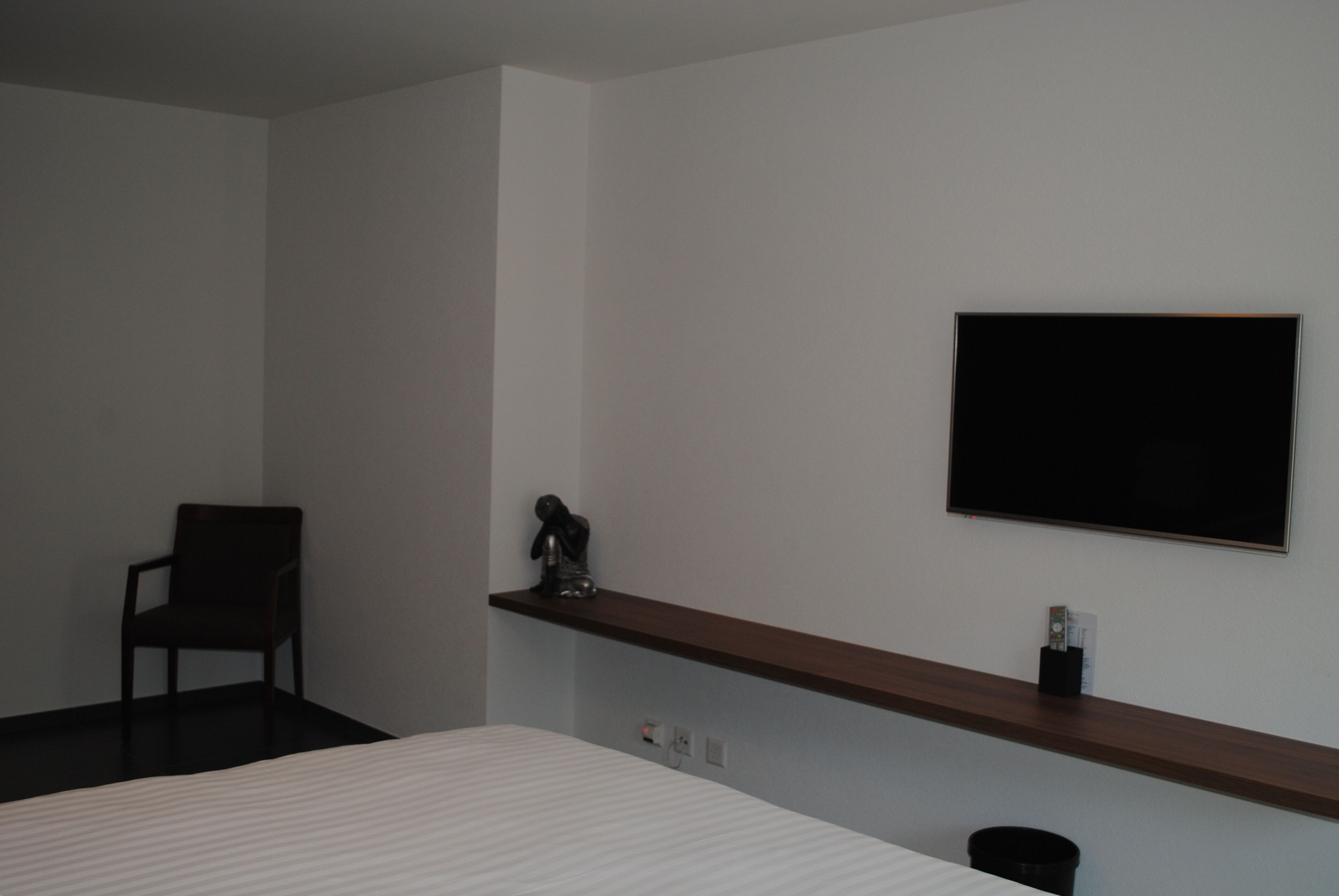 A hotel room with a bed, a chair, a shelf with a decorative item, and a flat-screen TV mounted on the wall.
