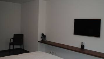A hotel room with a bed, a chair, a shelf with a decorative item, and a flat-screen TV mounted on the wall.