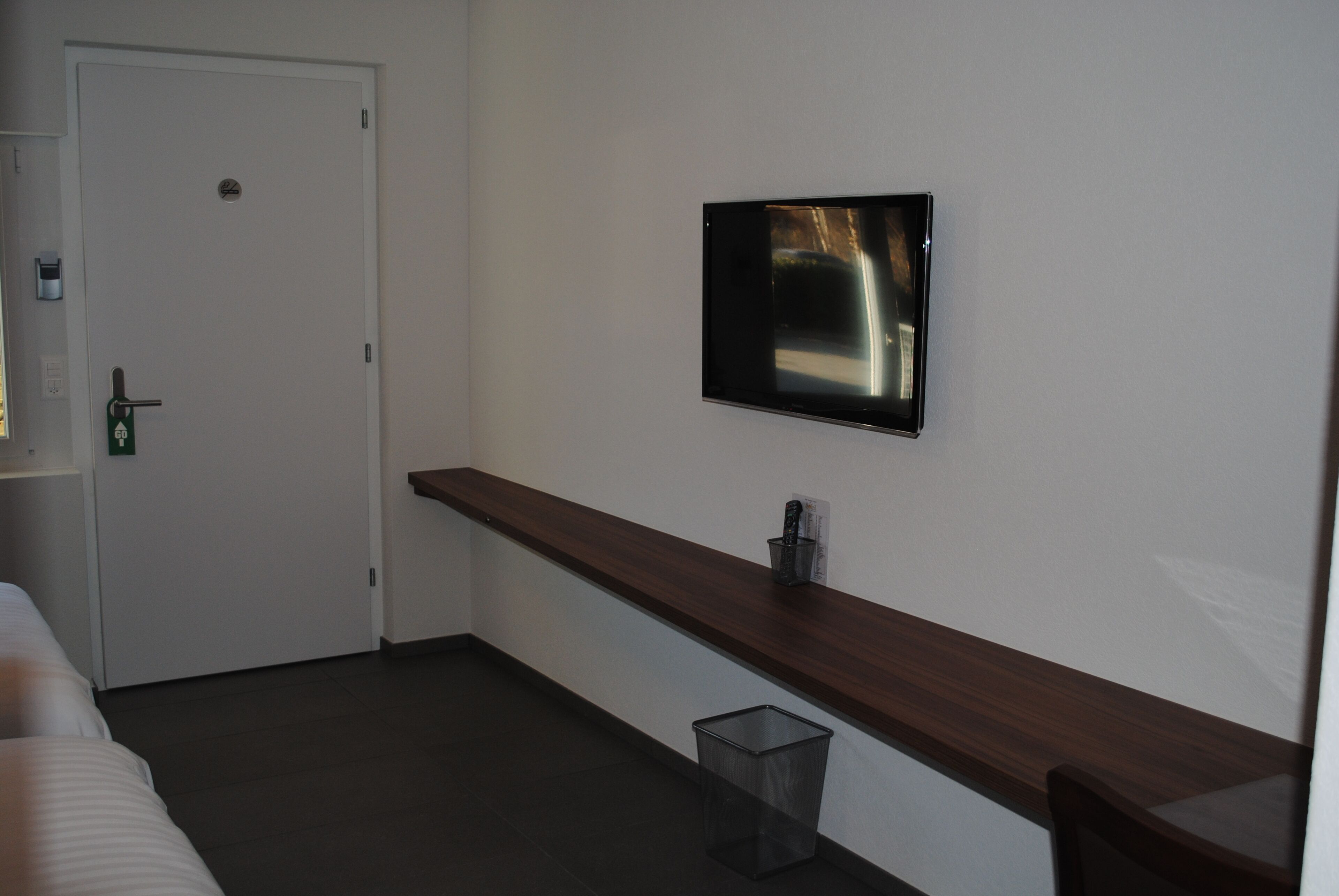 Una habitación de hotel con una cama, un banco de madera, un televisor montado en la pared y un cesto de basura.