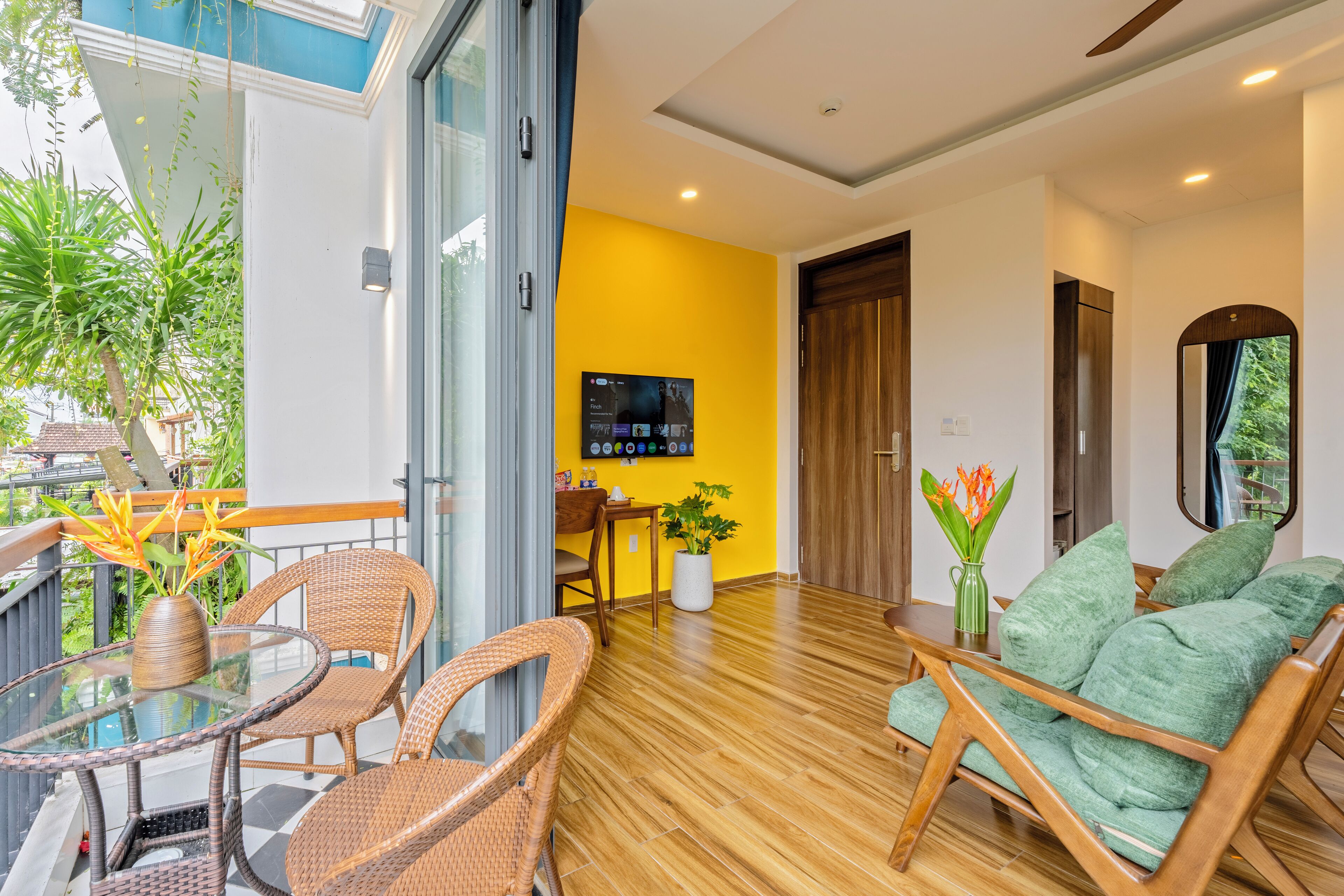 A modern living room with a wooden floor, a green sofa, wicker chairs, a glass table, and a TV mounted on the wall.