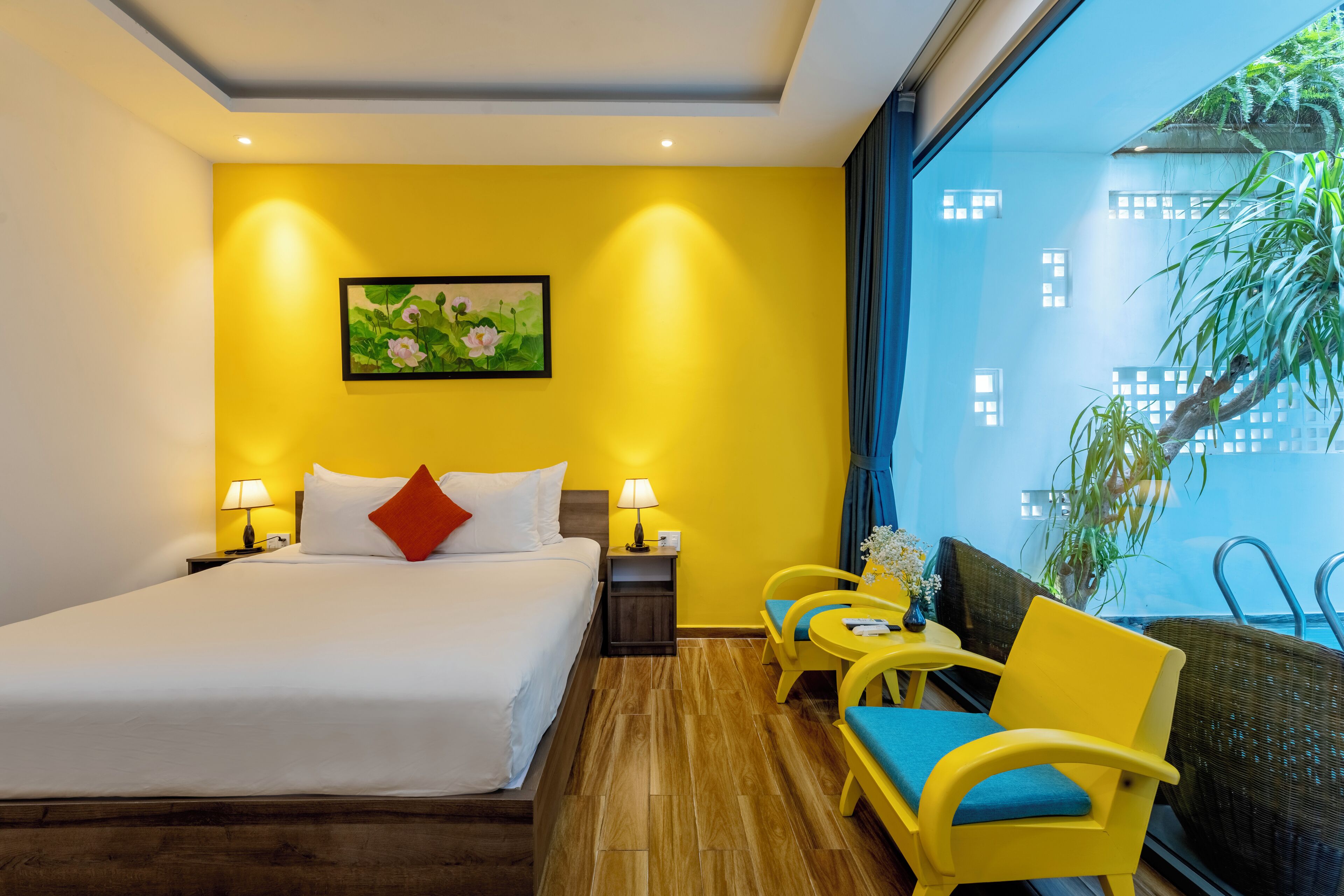 A hotel room with a bed, two bedside tables with lamps, a window with a view of greenery, and a seating area with yellow chairs.