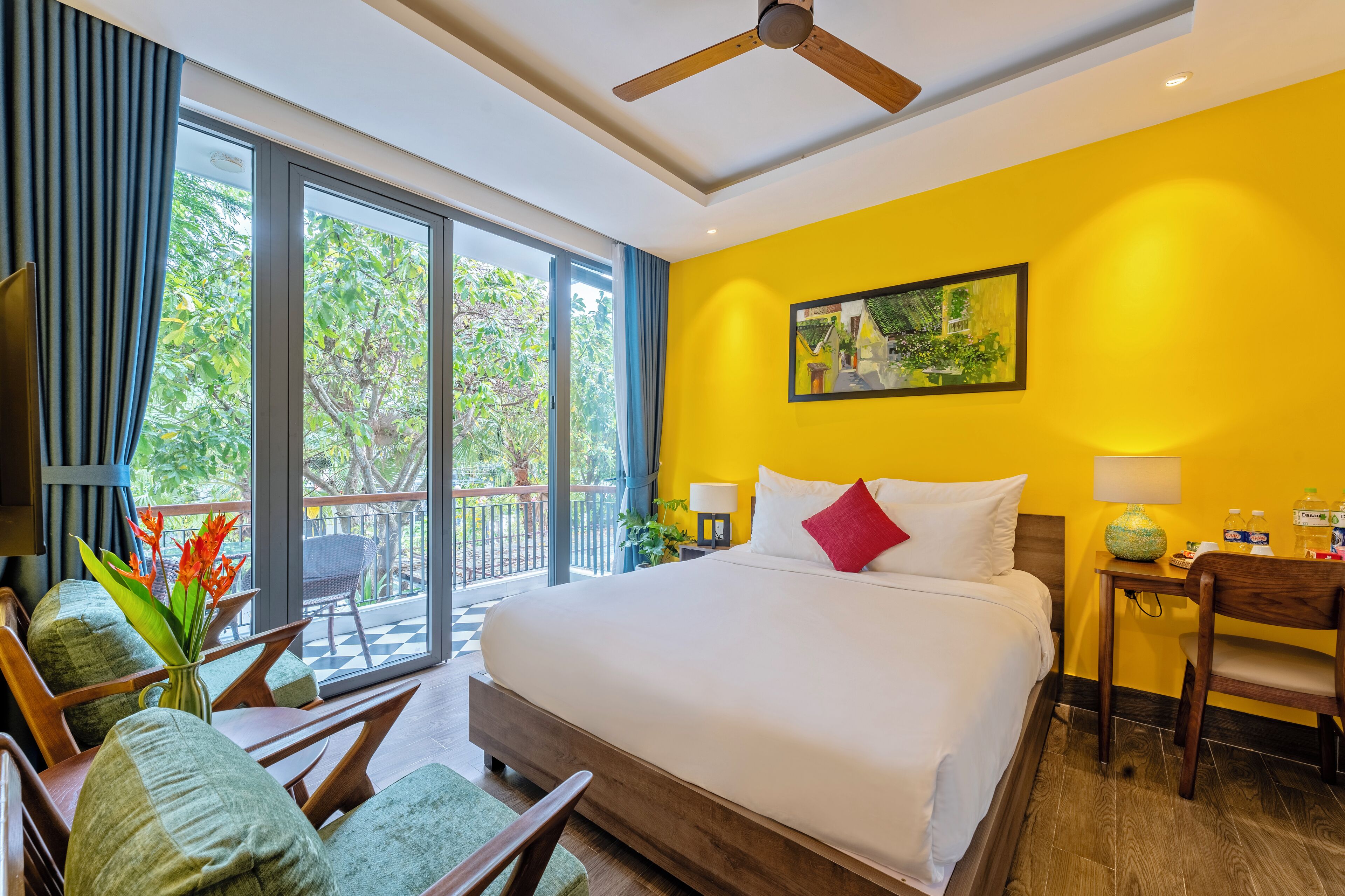 A hotel room with a large bed, a ceiling fan, a balcony with a view of trees, and a painting on the wall.
