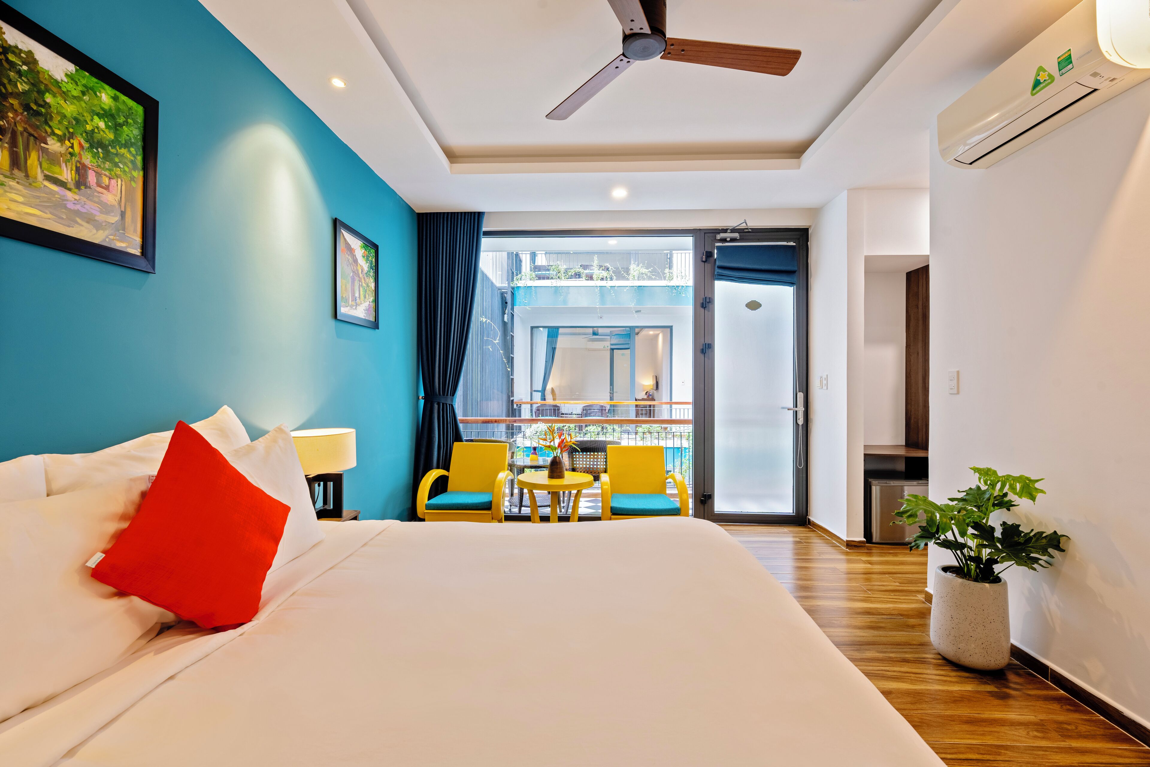 A hotel room with a bed, a ceiling fan, a balcony with yellow chairs, and a potted plant.