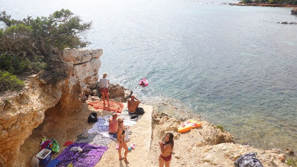Una playa rocosa con aguas claras, algunas personas y un barco a lo lejos.