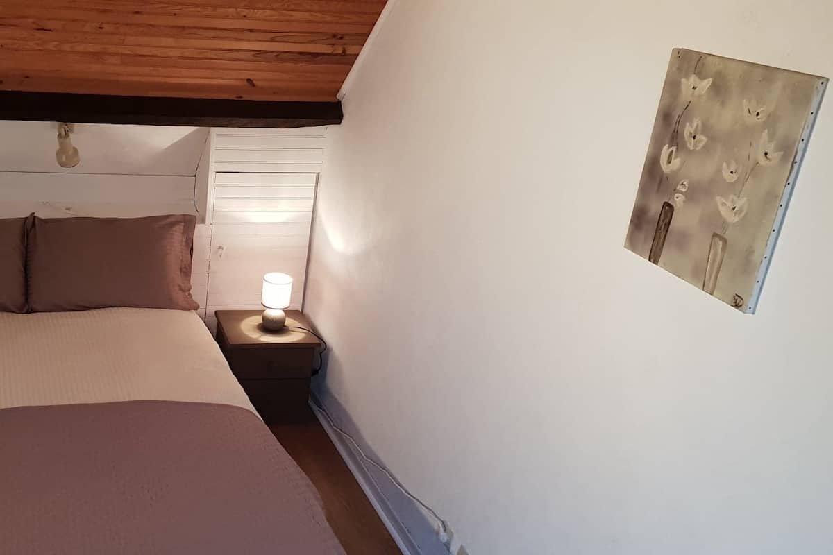 Un dormitorio con techo de madera, una cama con colcha color beige y marrón, dos mesitas de noche con lámparas y un aplique de pared.