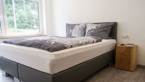 A modern bedroom with a bed, grey and white patterned bedding, a wooden nightstand, and a window with a view of greenery. - Hotel Haus am Berg (Pruemzurlay)