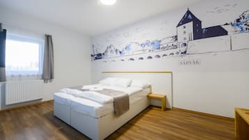 Een slaapkamer met een bed, een nachtkastje, een raam met gordijnen, een radiator en een muurschildering van een stadsgezicht met de naam 'SÁRVÁR'.