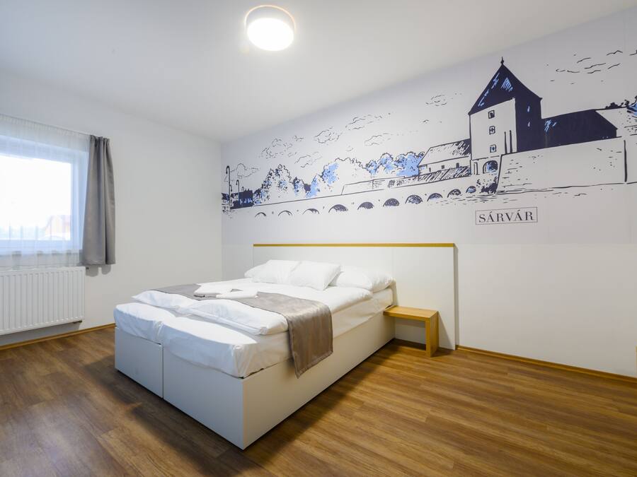 Een slaapkamer met een bed, een nachtkastje, een raam met gordijnen, een radiator en een muurschildering van een stadsgezicht met de naam 'SÁRVÁR'.
