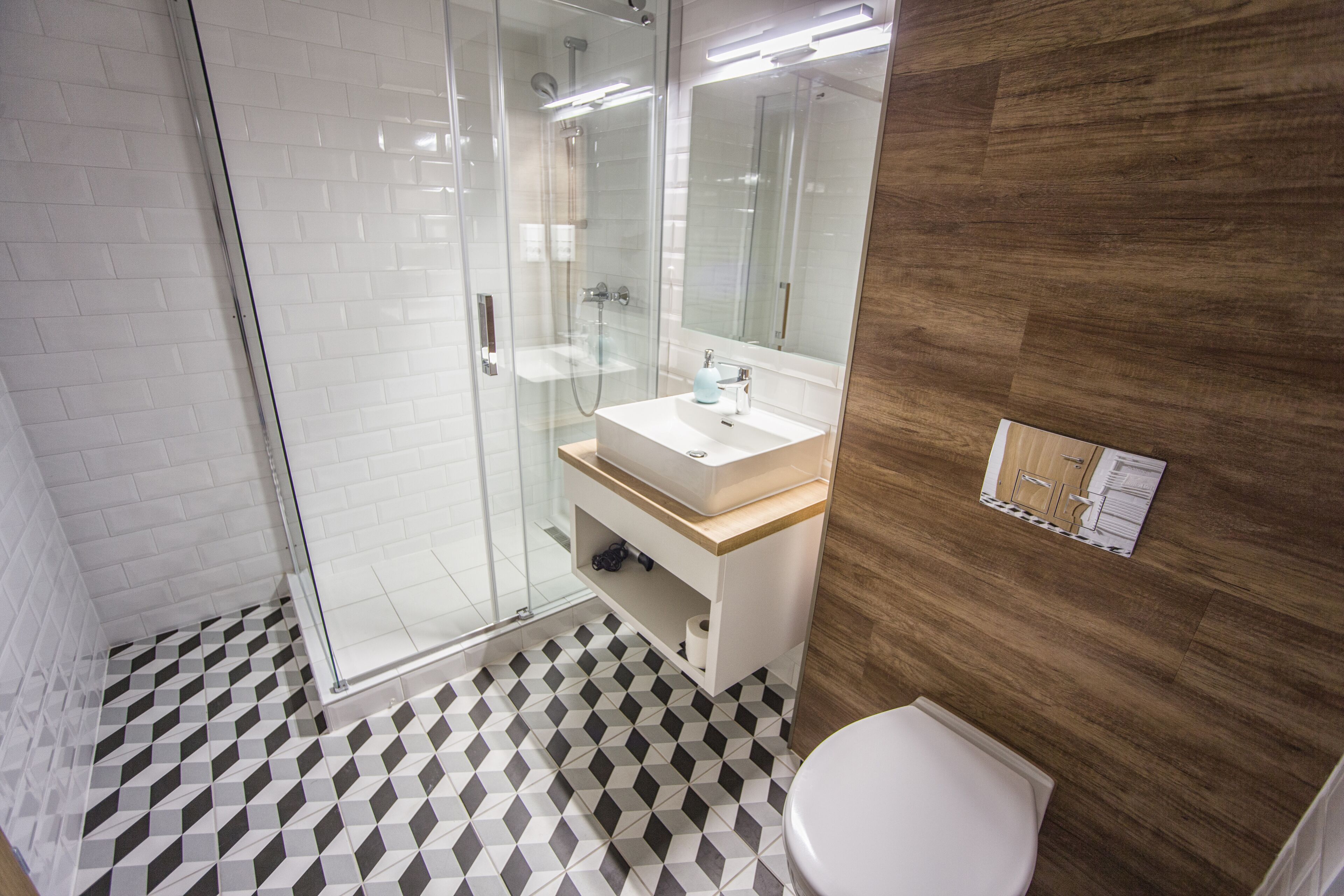 Un baño moderno con cabina de ducha de vidrio, un lavamanos blanco sobre encimera de madera y un suelo con estampado blanco y negro.