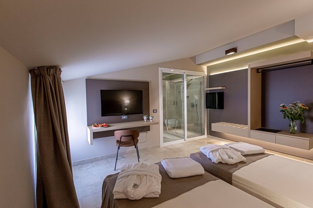 Habitación de hotel moderna con televisor de pantalla plana, cabina de ducha de vidrio, escritorio con silla y dos camas con toallas.