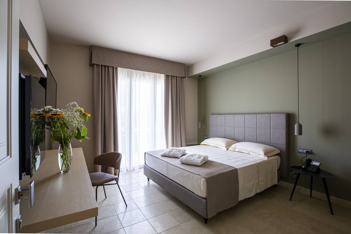 Habitación de hotel con cama, escritorio, silla, un florero con flores y un ventanal con cortinas.