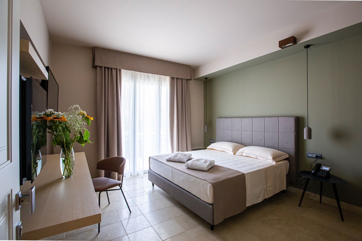 A hotel room with a bed, a desk with a chair, a vase with flowers, and a telephone.