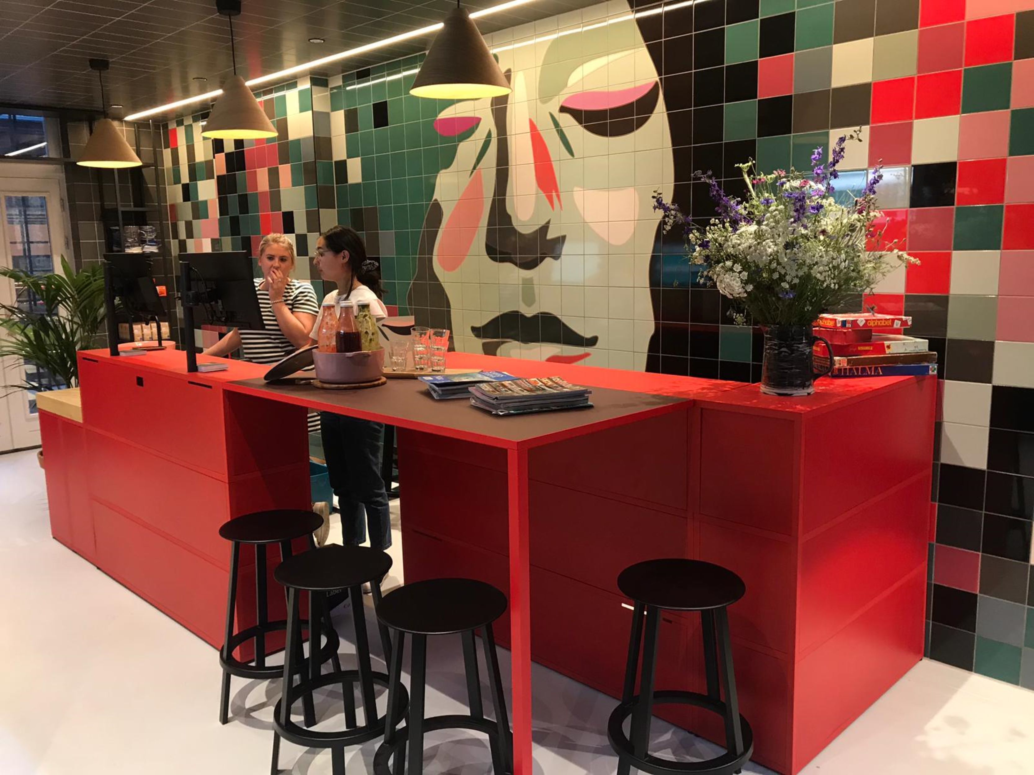Un mostrador de recepción rojo con taburetes negros, un gran mural abstracto y un jarrón con flores.