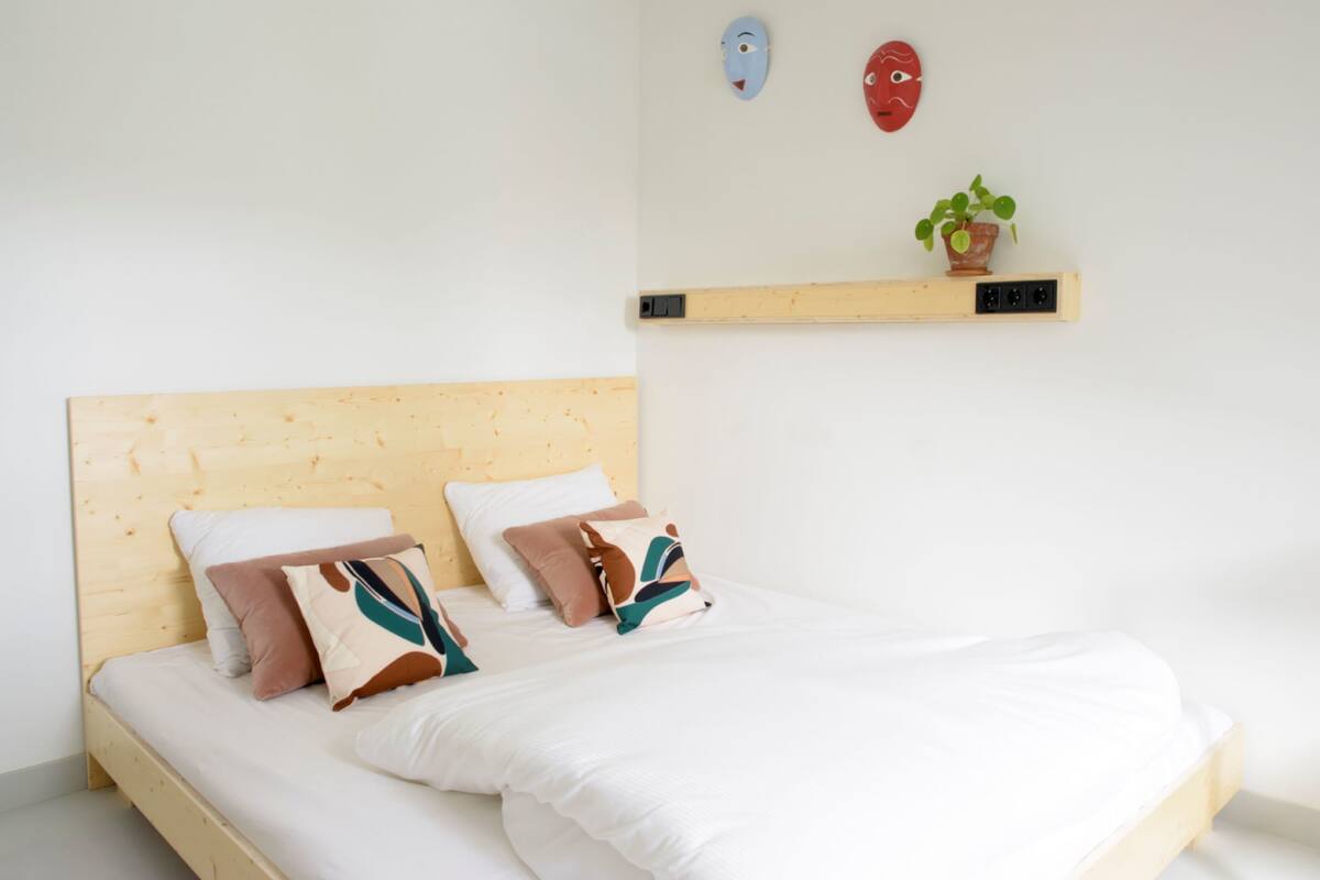 A bed with a wooden headboard, white bedding, and multiple pillows. There is a shelf with a plant and two masks on the wall above the bed.