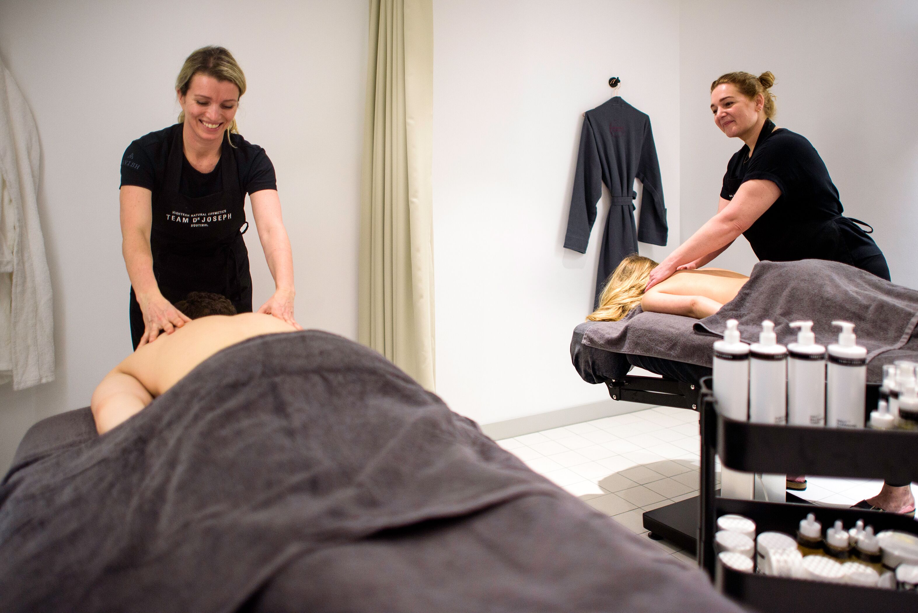 Ein Massageraum mit einer liegenden Person auf der Massageliege, zwei Therapeuten betreuen den Klienten, und ein Regal mit Massageölen und Lotionen.