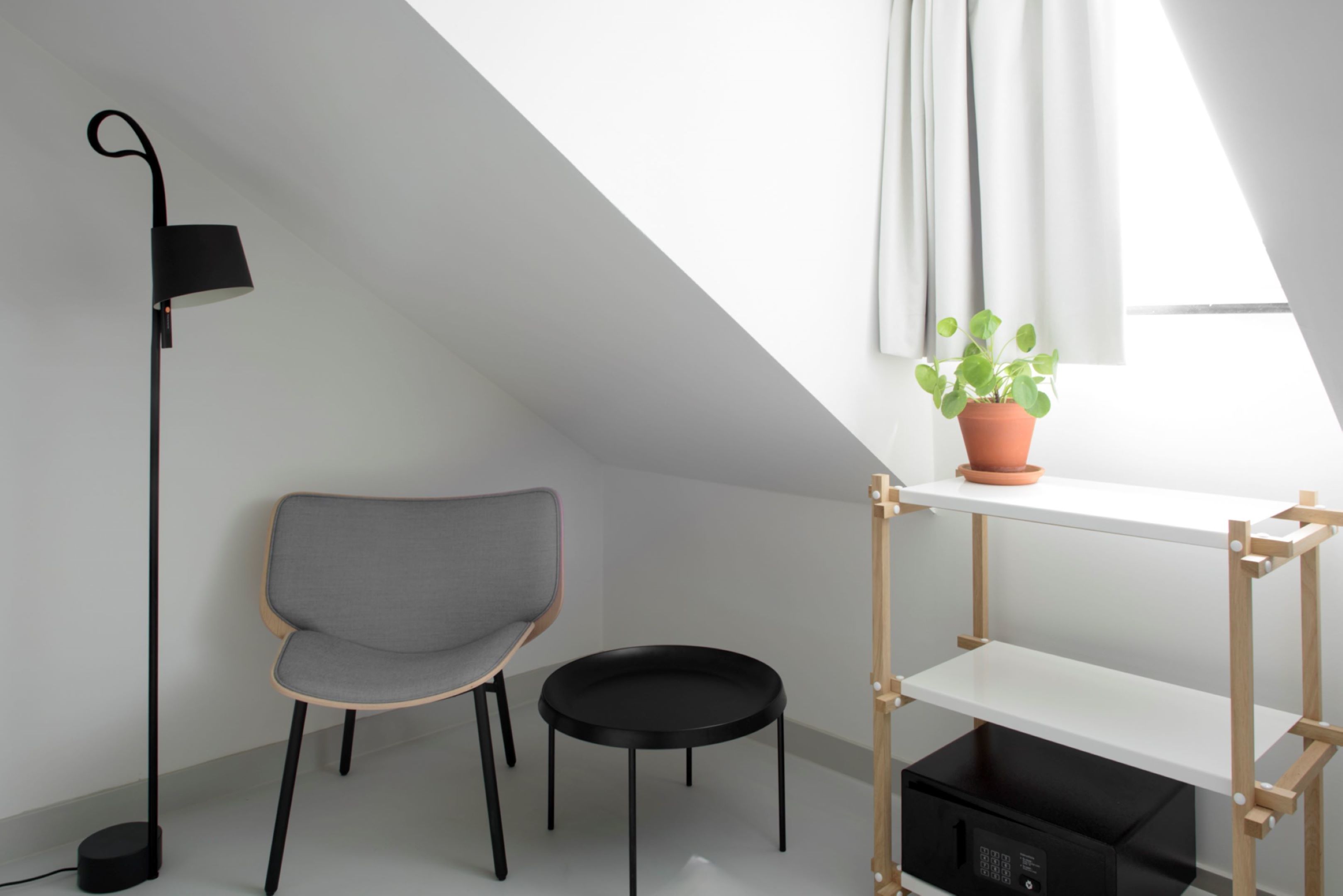 Une pièce moderne avec un fauteuil gris, un lampadaire noir, une petite table noire, une étagère en bois avec un pot de plante et une fenêtre avec des rideaux.