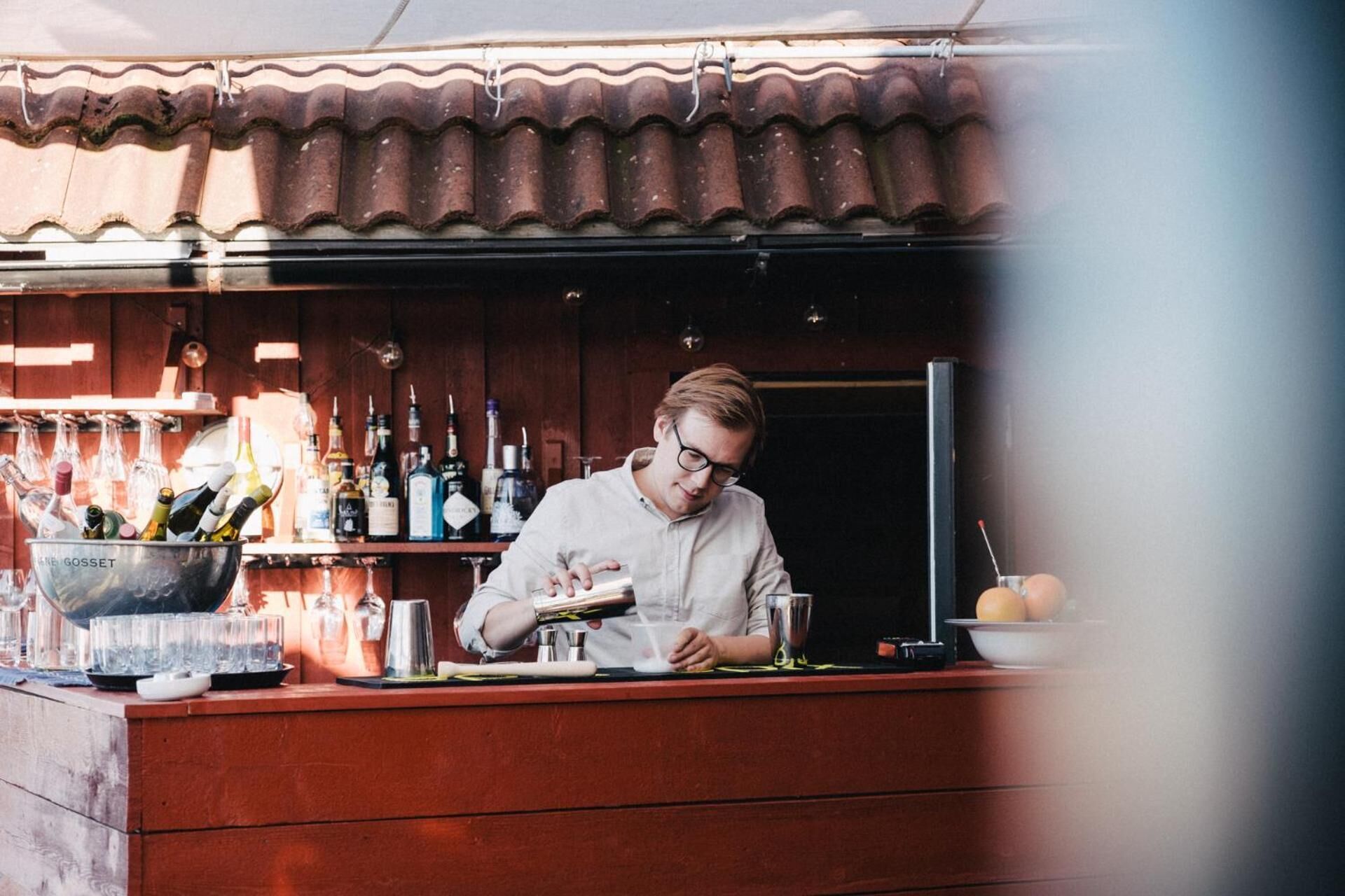 Een bar met een bartender die drankjes bereidt, diverse flessen en glazen op de planken, en een rustieke houten toonbank.