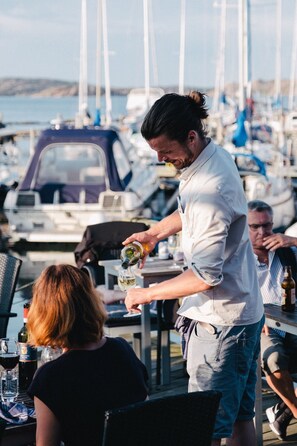 A person pouring a drink into a glass at a table with a view of boats in the background. - Björholmen Hotell & Restaurang (Klovedal)
