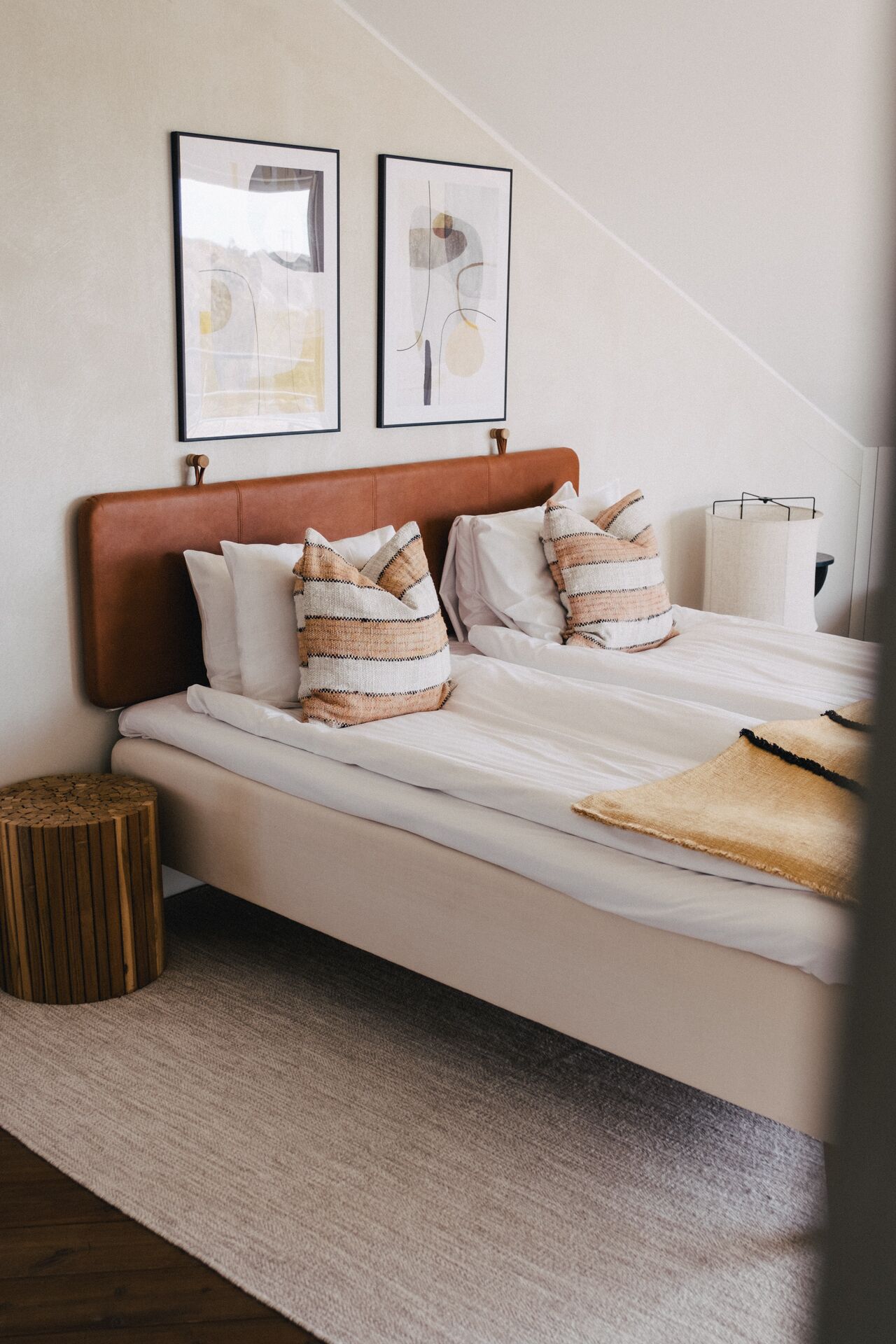Un lit bien fait, avec une tête de lit marron, une literie blanche et des coussins à rayures. Deux œuvres d’art encadrées sont accrochées au-dessus du lit, un tabouret en osier se trouve à côté, et une lampe est posée sur la droite.