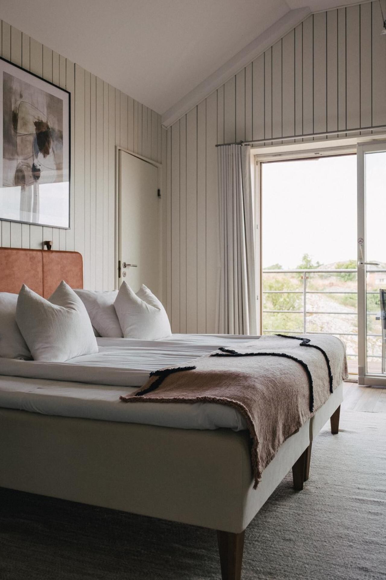 Un dormitorio con una cama grande, ropa de cama blanca, un edredón marrón y vistas a zonas verdes a través de un gran ventanal.