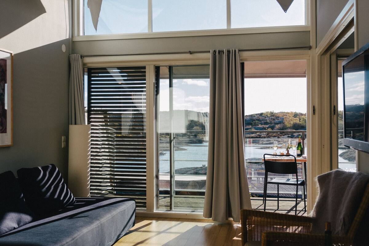 Un salon moderne avec un canapé, un fauteuil et une table sur laquelle se trouvent une bouteille et des verres. Une grande porte-fenêtre coulissante donne accès à un balcon d’où l’on peut admirer un lac et des collines.