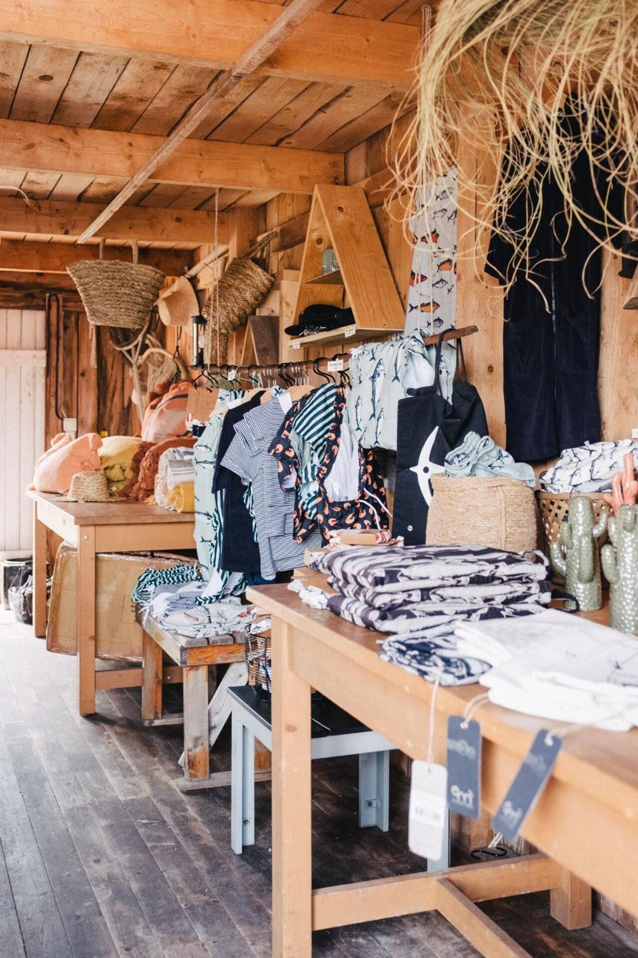 Een rustiek houten interieur met tafels waarop verschillende textiel- en kledingstukken staan uitgestald.