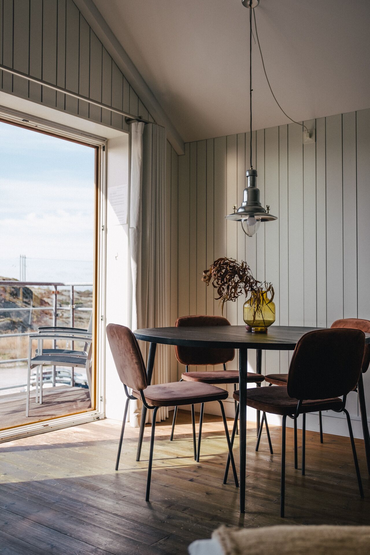 A dining area with a table and chairs, a hanging light fixture, and a view through a large window.