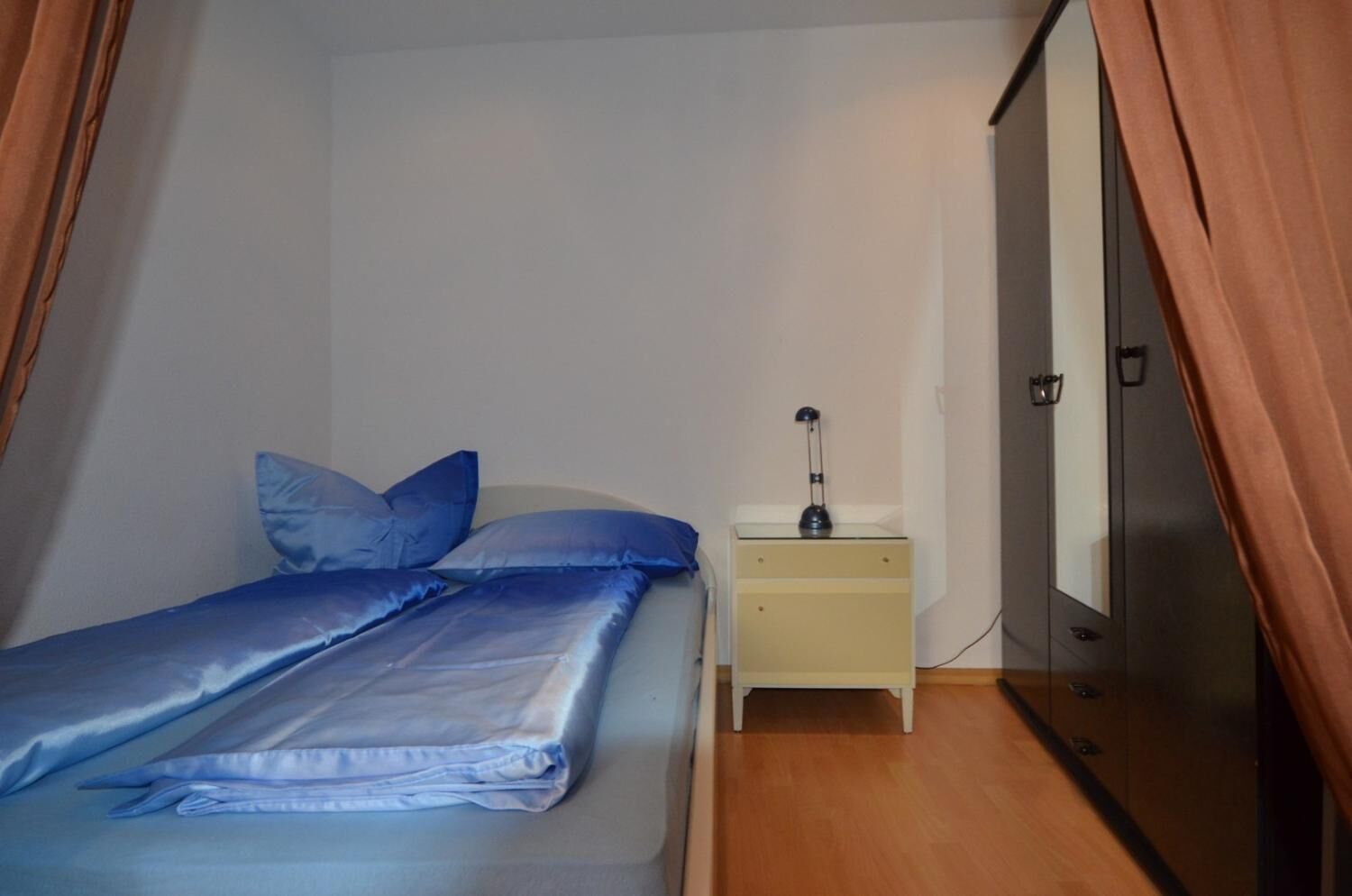 A small, single-bed room with a wooden floor, a nightstand with a lamp, and a wardrobe.