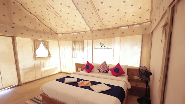 Un dormitorio amplio con una cama grande, varias almohadas, un ventilador de pie y una ventana con cortinas.